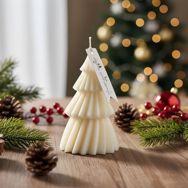 White Christmas tree-shaped candle with a decorative tag on a wooden surface surrounded by pinecones, red ornaments, and.