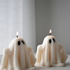 Two white ghost-shaped candles with black eyes and lit wicks on a light surface.