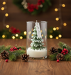 Glass jar candle with a detailed green pine tree shaped wax inside on wooden table with pinecones and red berries.