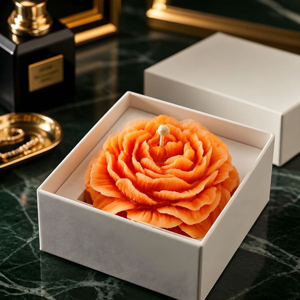 Large handmade candle shaped like an orange peony flower in a white gift box on dark green marble surface.