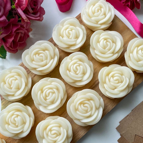 Set of twelve white rose-shaped candles arranged neatly on a wooden tray with pink roses and a pink ribbon nearby.