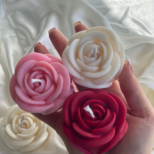 Hand holding four medium rose-shaped candles in white, pink, cream, and dark red colors.