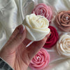Hand holding a white rose-shaped candle with various pink and red rose candles on white fabric background.
