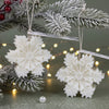 White snowflake-shaped candles hanging on frosted pine branches with warm fairy lights background.