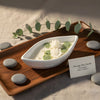 White ceramic boat-shaped candle with green accents and floral wax details on wooden tray.
