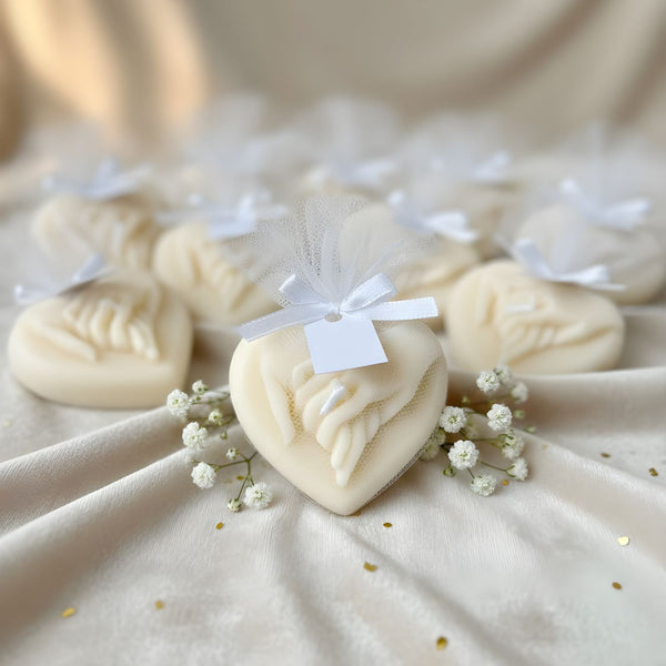 Cream-colored heart-shaped candle with intertwined hands design, tied with white ribbon bow and small tag, surrounded by.