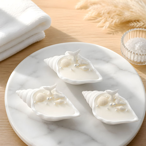 Three white conch shell soy wax candles with small decorative seashells on top, arranged on a round marble tray.
