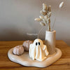 White ceramic Halloween ghost candle with black eyes on beige wavy tray with dried flowers and small pumpkins.