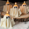 Four small white candles shaped like ghosts with black oval eyes, lit and placed on a wooden surface.