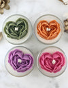 Four glass candles with 3D heart-shaped flowers in green, orange, purple, and pink on a marble surface.