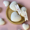 Set of white heart-shaped candles with textured surfaces displayed on a round wooden plate and pink background.