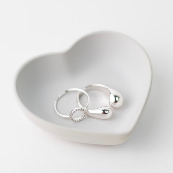 White ceramic heart-shaped tray holding two silver rings and a silver heart pendant.
