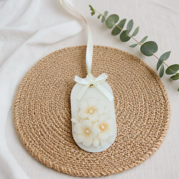 White oval soy wax sachet with delicate cream flowers, tied with a white ribbon bow, on woven mat.