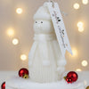 White snowman candle wearing knit hat and scarf with Merry Christmas tag and red ornaments on snow-like surface.