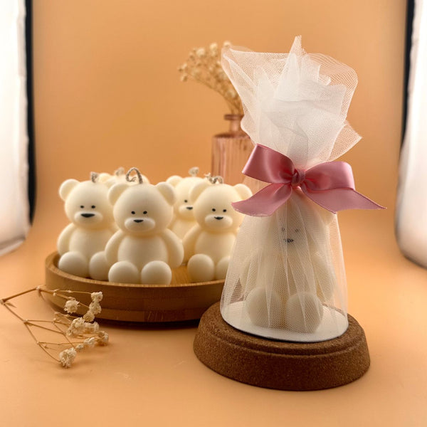 White teddy bear-shaped candle with round features, tied with pink ribbon in translucent wrapping on wooden base.