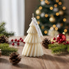 White Christmas tree-shaped candle with a decorative tag on a wooden surface surrounded by pinecones, red ornaments, and.