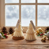 Two cream-colored Christmas tree-shaped candles with wooden tags on a wooden table, surrounded by holiday decor and blurred.