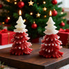 Two red and white layered pine tree candles with lit wicks on a wooden surface, Christmas tree and gifts blurred in.