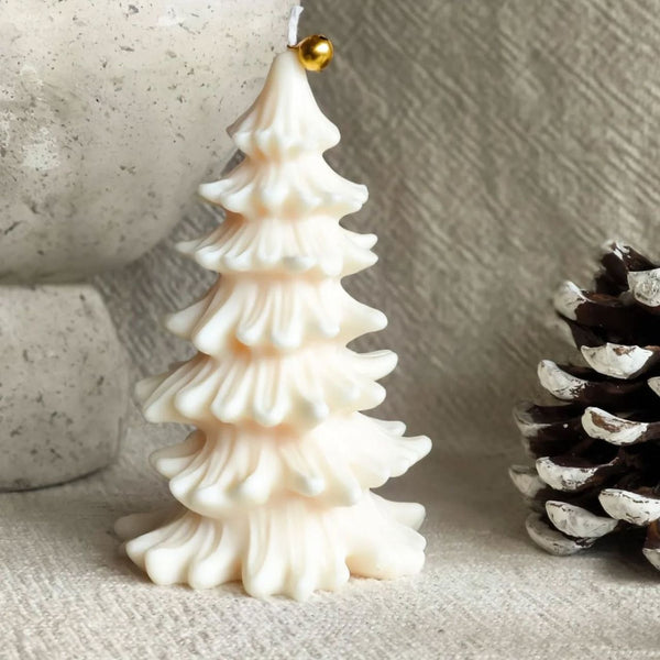 White pine tree-shaped candle with gold ornament on textured beige surface.