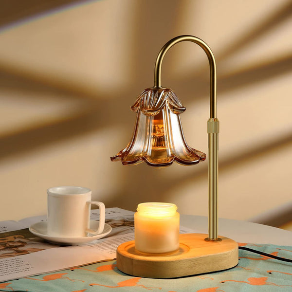 Electric candle warmer lamp with brass curved arm and amber glass flower-shaped shade over lit candle on wooden base.