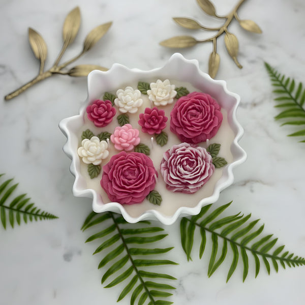 White scalloped candle bowl filled with pink and white floral wax candles on marble surface with green fern leaves.