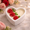 White heart-shaped ceramic candle jar with red rose and green leaf wax decorations on cream wax surface.