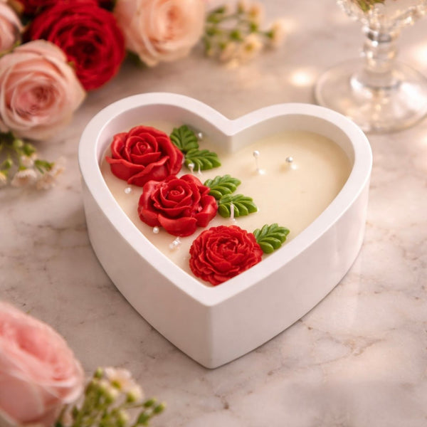 White heart-shaped ceramic candle jar with red rose and green leaf wax decorations on cream wax surface.