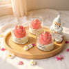 Three glass jar candles with pink lotus flower wax tops on a round wooden tray, next to a small white Santa figurine.