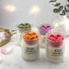 Set of four glass jar candles with 3D heart-shaped flowers in orange, pink, purple, and green on white surface.