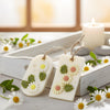 Two white soy wax hanging sachets embossed with green leaves and daisy flowers, tied with natural twine.
