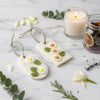 Two white soy wax hanging sachets decorated with dried flowers and leaves, each with a twine loop for hanging, displayed on.