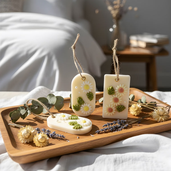 Two white soy wax hanging sachets with embedded green and orange dried flowers on a wooden tray with dried lavender and.
