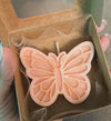 Peach-colored butterfly-shaped candle with detailed wing patterns in a brown gift box with mesh lining.