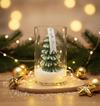 Glass jar candle with a green pine tree-shaped wax inside, surrounded by golden ornaments and warm fairy lights.