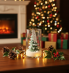 Clear glass jar candle with a green pine tree-shaped wax center and white base, surrounded by pine cones, holly leaves, and.