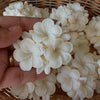 Hand holding a white flower-shaped aromatic candle with multiple petals above a basket filled with similar candles.
