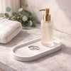White oval ceramic tray holding a clear pump bottle and two silver rings on marble surface.