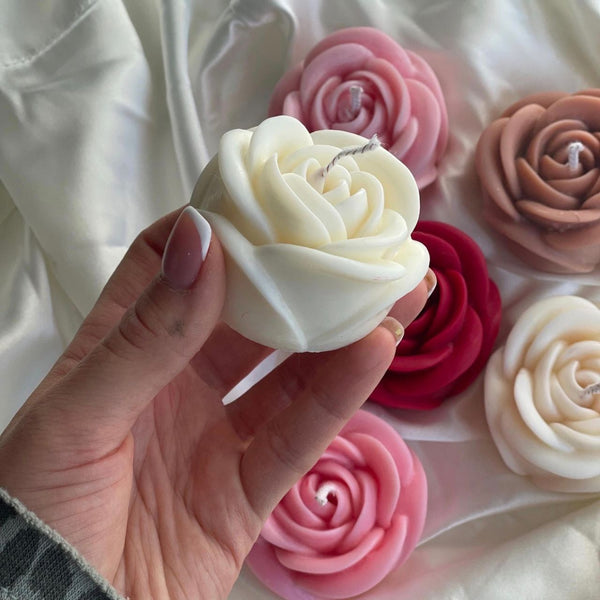 Hand holding a white rose-shaped candle with various pink and red rose candles on white fabric background.