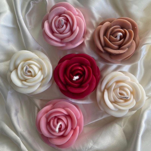 Six medium rose flower candles in pink, red, cream, and beige shades arranged on white satin fabric.