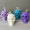 Four Halloween skull candles in purple, cream, teal, and violet with detailed bone texture and twisted wicks.