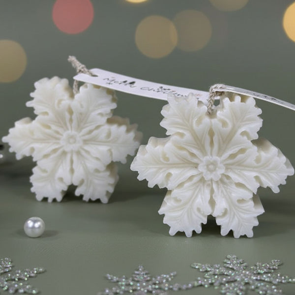 Two white snowflake-shaped candles with intricate details on a green surface, surrounded by silver snowflake decorations and .