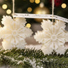White snowflake-shaped candles hanging from rustic twine with a festive background and pine branches below.