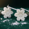 White snowflake-shaped candles with detailed patterns on a dark green surface with scattered faux snow and pearls.