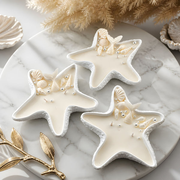 Three white handmade starfish-shaped candles with embedded seashells on a round marble tray.