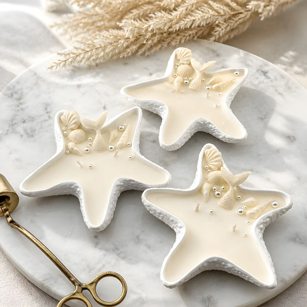 Creamy white starfish-shaped candles with seashell accents on matching starfish trays on marble surface.