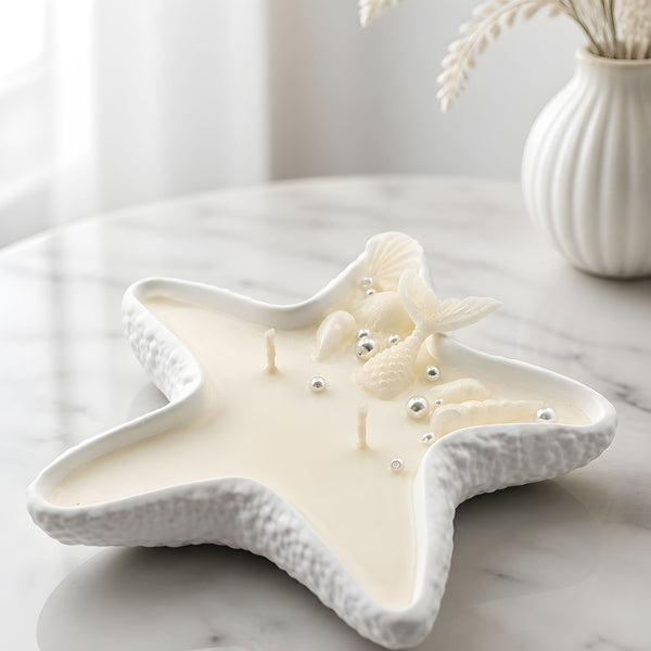 White handmade starfish-shaped candle with embedded seashells on a marble table.