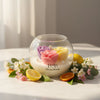 Round glass jar candle with pastel rose-shaped wax melts in pink, purple, and yellow, surrounded by citrus fruits and white.