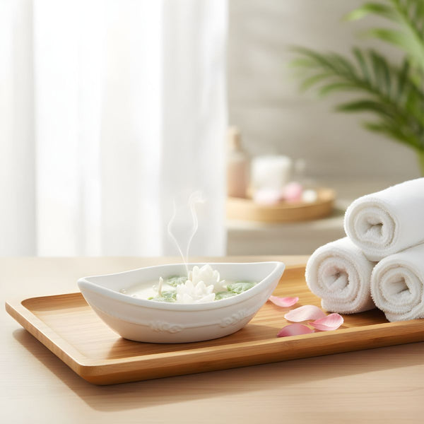 White ceramic bowl candle with flower petals on wooden tray beside rolled white towels and green plant.