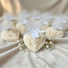 Cream-colored heart-shaped candle with intertwined hands design, tied with white ribbon bow and small tag, surrounded by.