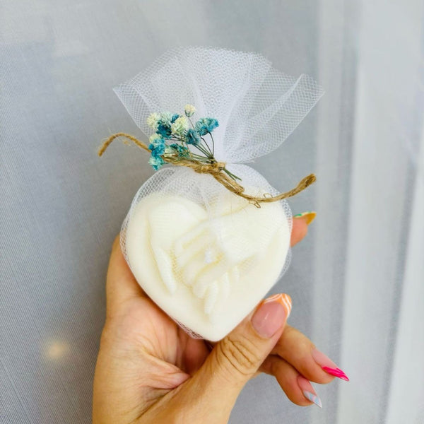 White heart-shaped candle with intertwined hands design, wrapped in sheer white fabric and tied with twine and small blue.